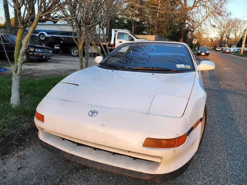 White Toyota MR2 sports car with its headlights down, parked beside the road